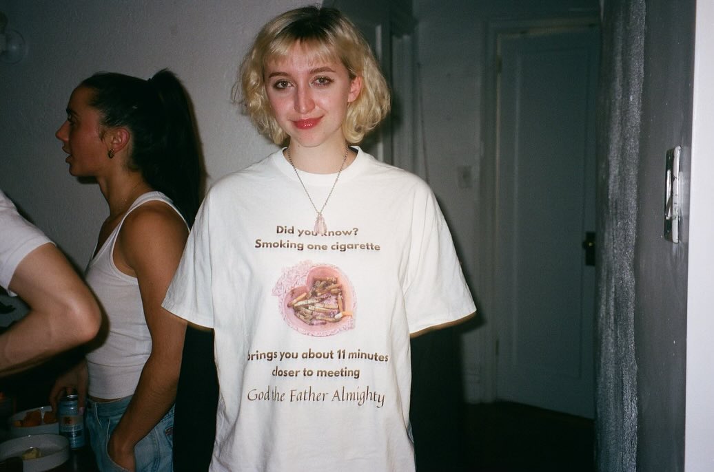 A person standing indoors wearing an oversized white graphic T-shirt with printed text and a circular illustration on the front. Another person is partially visible in the background near a mirror.