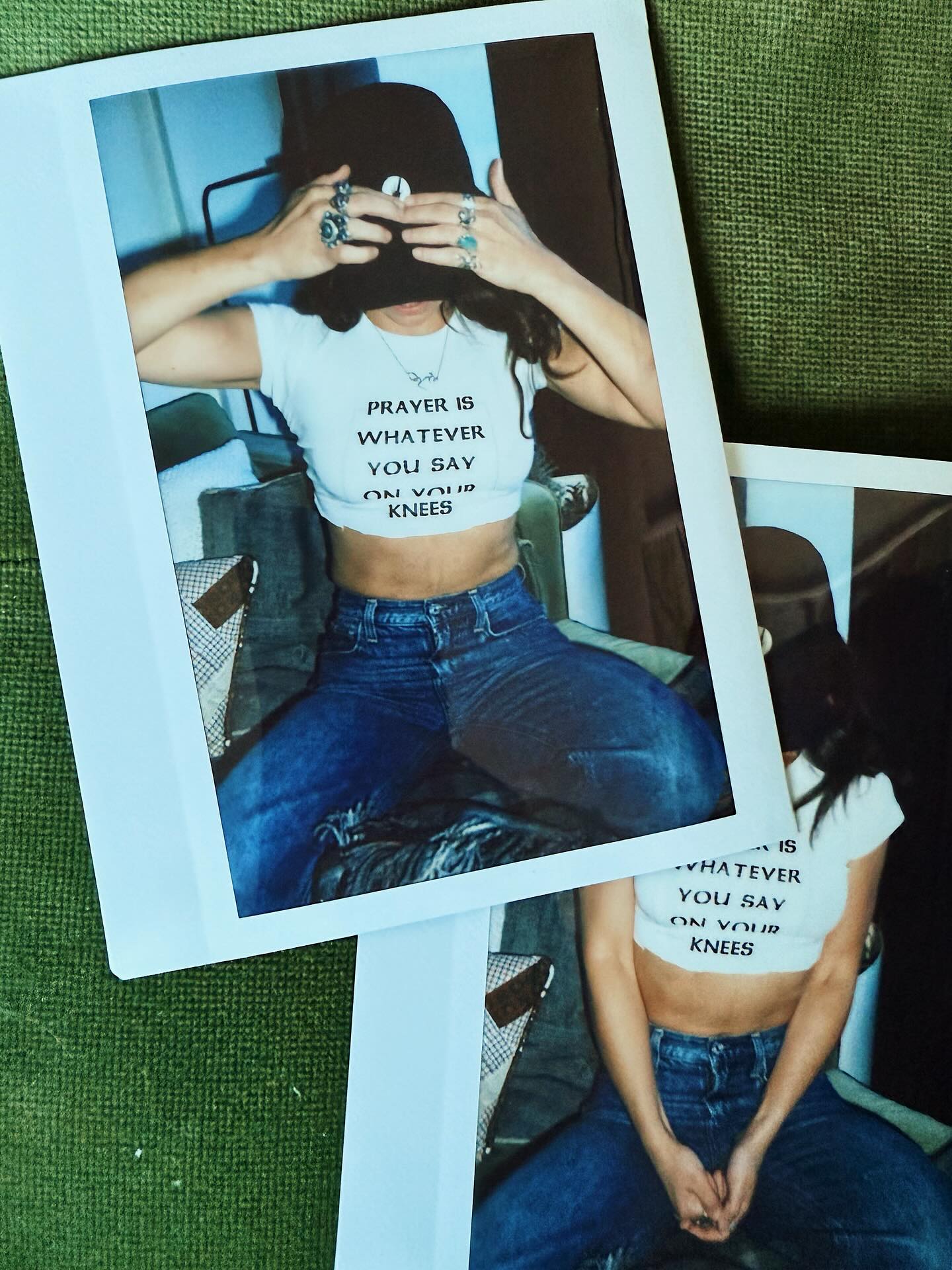 A printed instant-film-style photo print showing a person wearing a white graphic crop top with black text and blue jeans, placed on a textured green surface with additional overlapping prints visible.