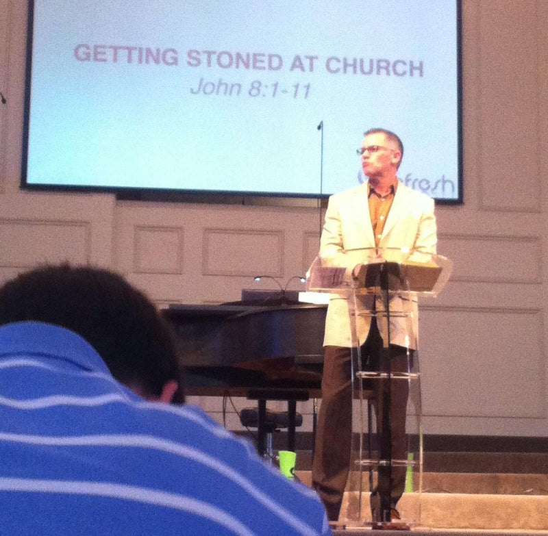 Getting Stoned at Church, John 8:1-11. A pastor preaches at a clear podium in front of a projector screen showing the sermon title.