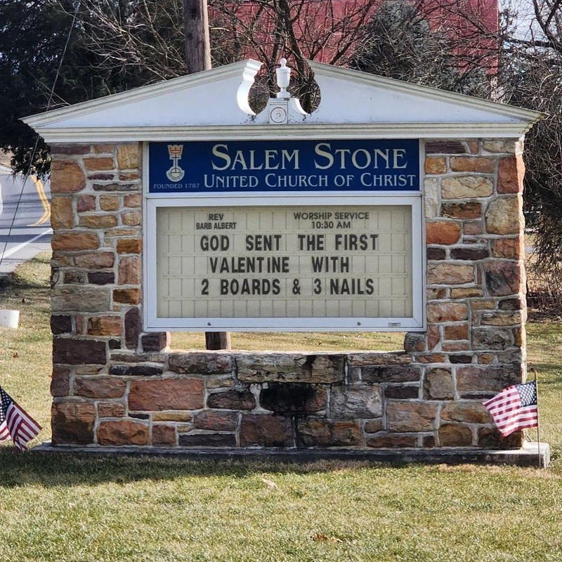 Church sign: God sent the first valentine with 2 boards & 3 nails. A Salem Stone United Church of Christ marquee sign flanked by American flags.