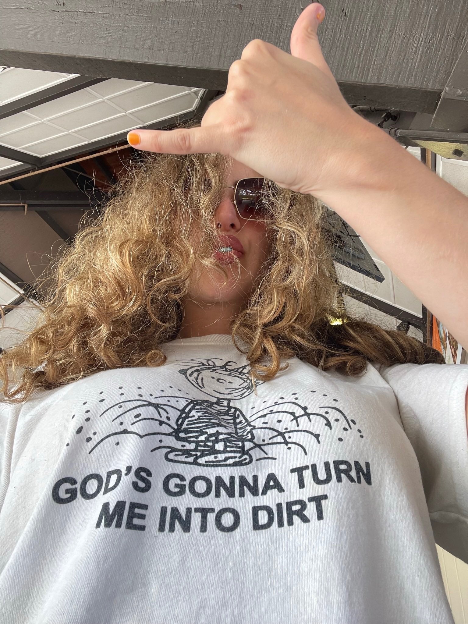 A person wearing a white graphic t-shirt with a black printed design and the text 'GOD'S GONNA TURN ME INTO DIRT', taking a selfie with a hand gesture.