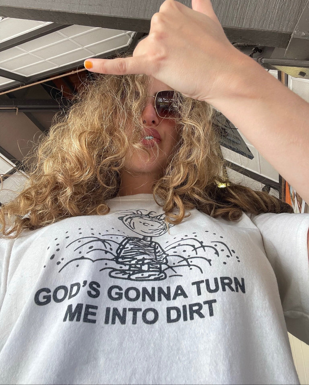 A person wearing a white graphic t-shirt with a black printed design and the text 'GOD'S GONNA TURN ME INTO DIRT', taking a selfie with a hand gesture.