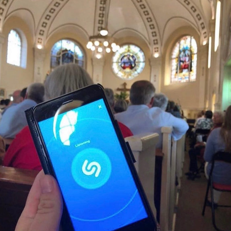 A phone held up in a church pew with Shazam open and Listening, trying to identify the hymn during a service in front of stained glass windows.
